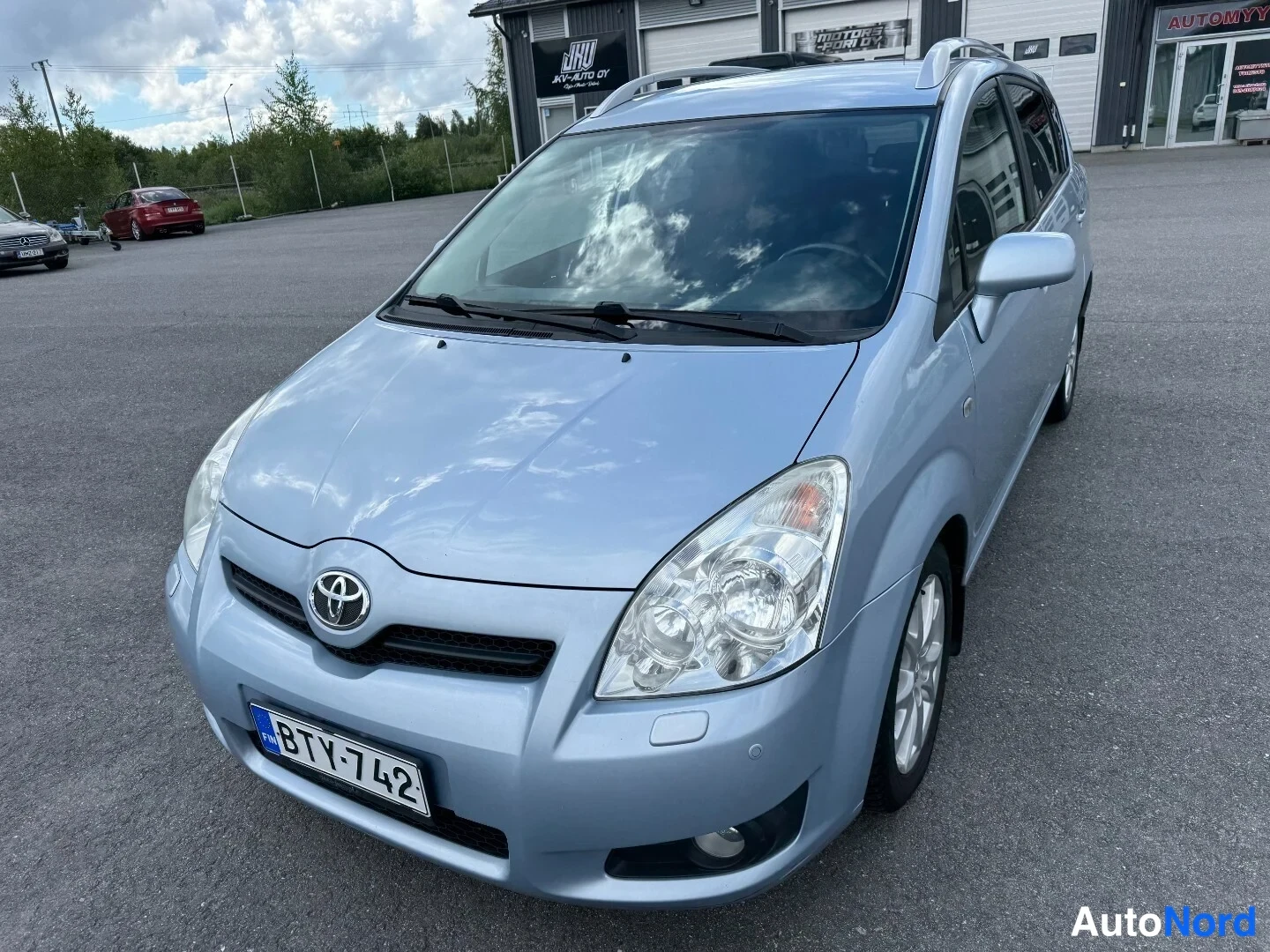 Etuviisto, nokka vasemmalle: Vaaleansinisen Toyota Corolla Verson etuviisto vasemmalta, rekisteritunnus, kattokaiteet näkyvät.