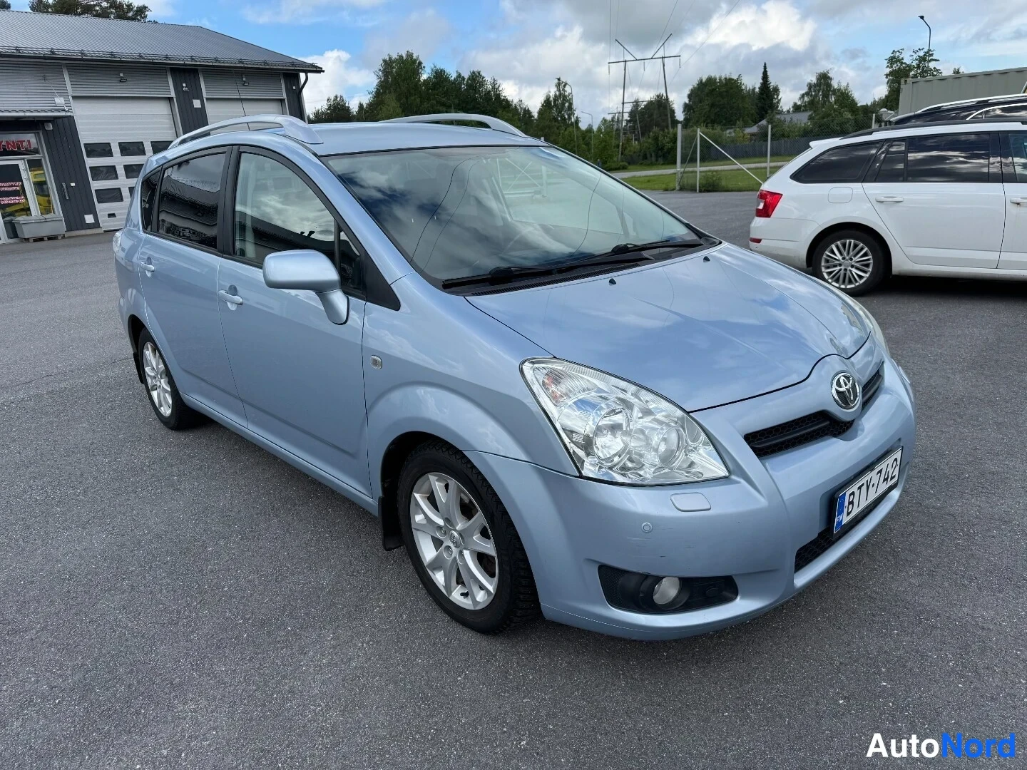 Etuviisto, nokka oikealle: Toyota Corolla Verso -henkilöauto vasemmasta etukulmasta, vaaleansininen koriväri, kattokaiteet ja hopeanväriset vanteet.
