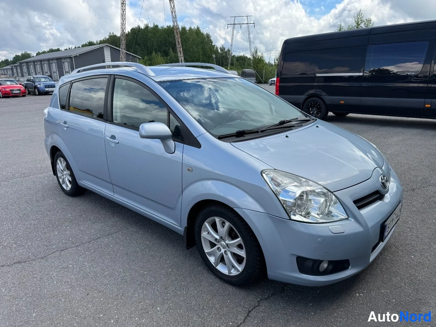 Etuviisto, nokka oikealle: Toyota Corolla Verso -henkilöauto vasemmasta etukulmasta, vaaleansininen koriväri, kattokaiteet ja viisiovinen kori.