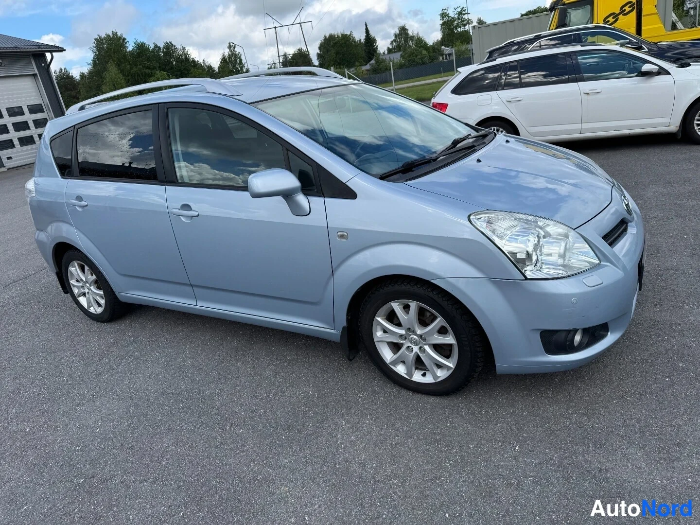 Etuviisto, nokka oikealle: Toyota Corolla Verso -henkilöauto vasemmasta etukulmasta, vaaleansininen koriväri, kattokaiteet ja hopeanväriset vanteet.