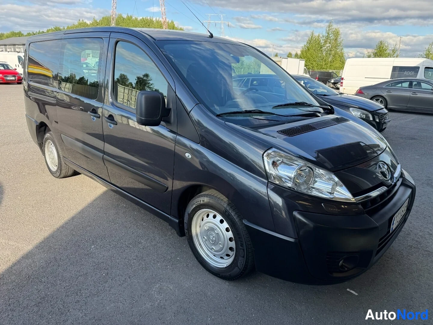 Etuviisto, nokka oikealle: Toyota Proace -pakettiauto tummanharmaana kolmanneltakulmalta edestä oikealta, pyörivät pyörät ja tarrat pyöränkaarella.