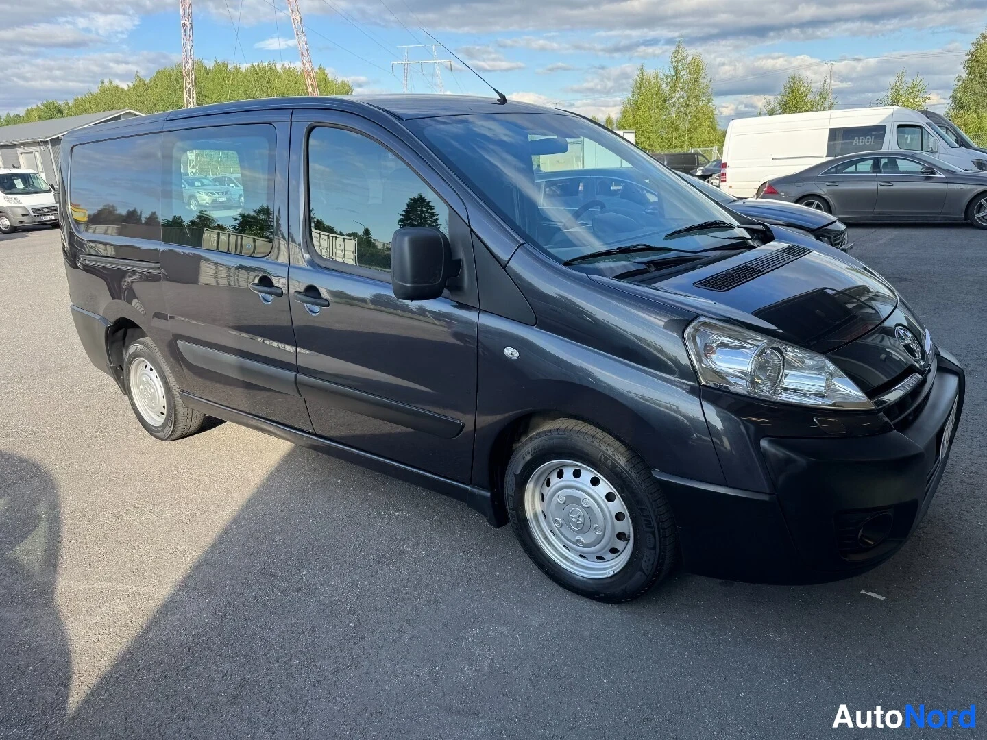 Etuviisto, nokka oikealle: Tummanharmaa Toyota Proace -pakettiauto etuviistosta oikealta, sivuovet ja etuosa kuvassa, teräsvanteet pienillä muovitöpsleillä.