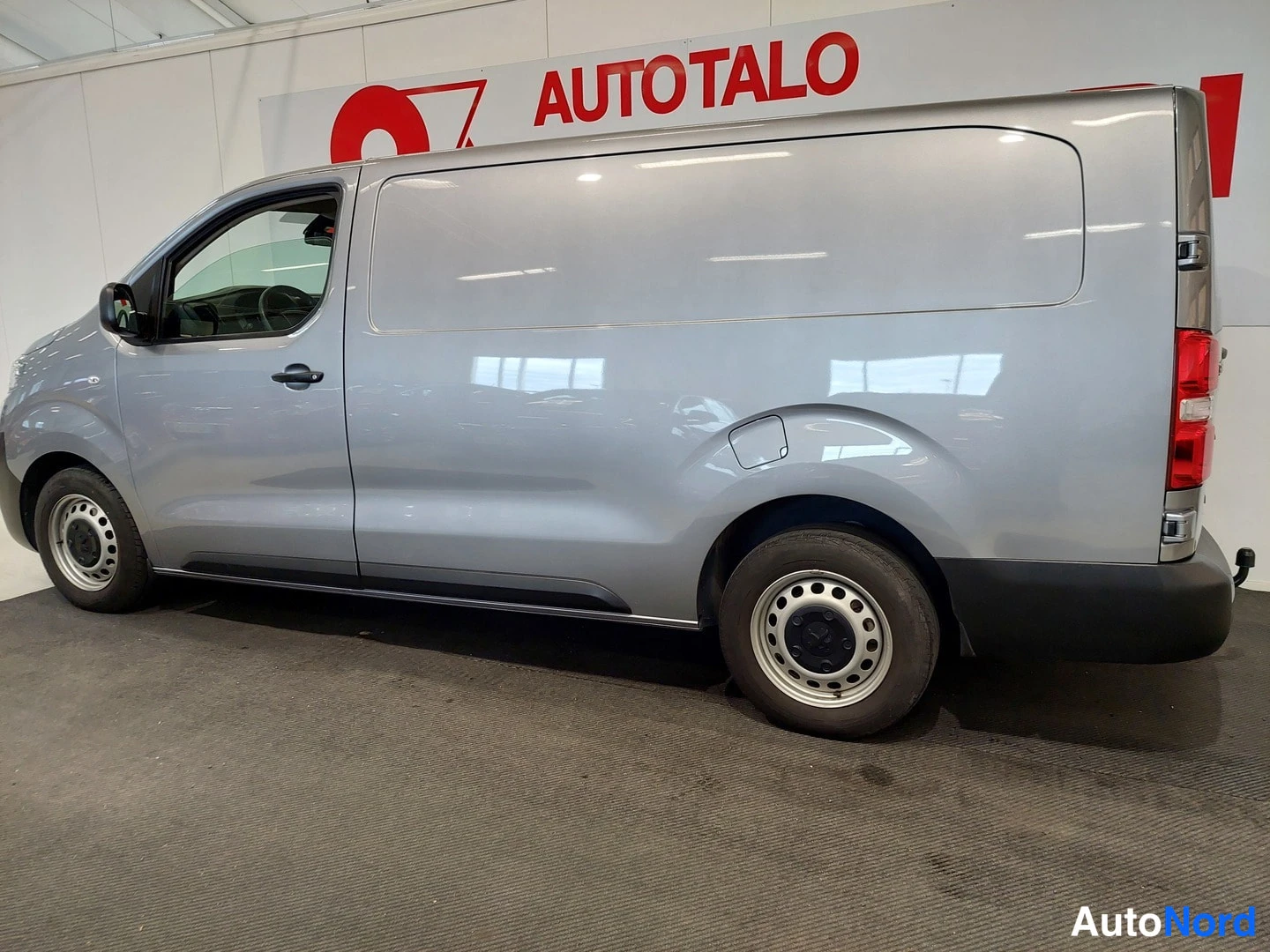 Takaviisto: Pakettiauto sivulta takaa, hopeanharmaa kori, lattiaan aukeava liukunäyttö 없는 sivupaneeli, pyöränkannen alla tumma muovilista.