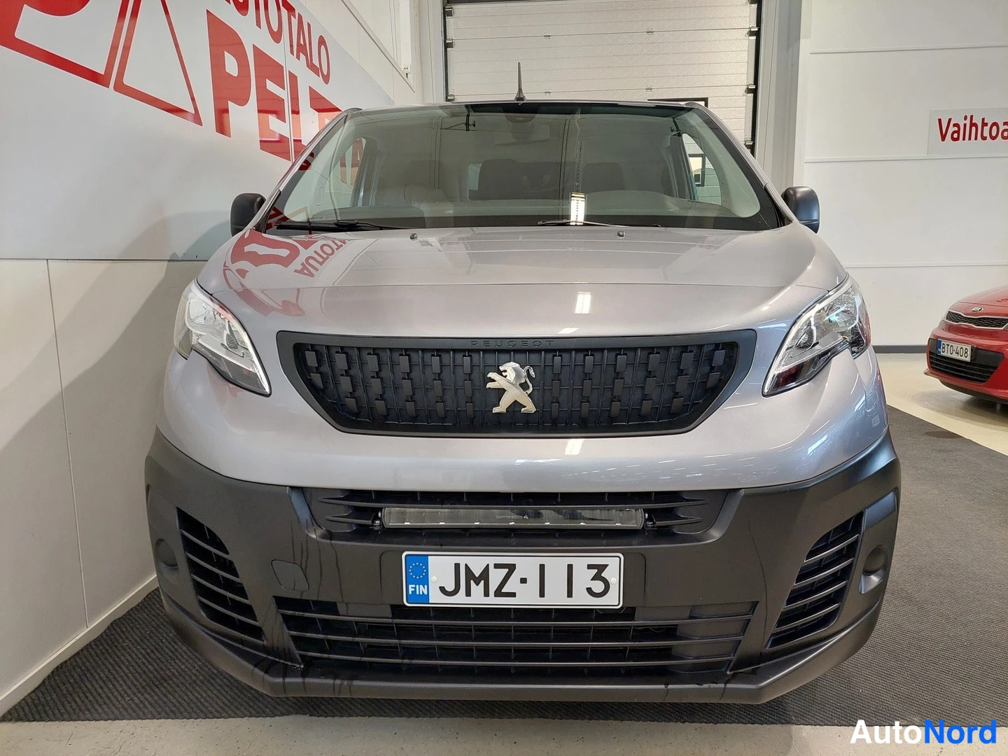 Edestä: Peugeot-pakettiauto suoraan edestä, musta säleikkö leijonamerkillä, sumuvalot puskurissa, rekisteri.