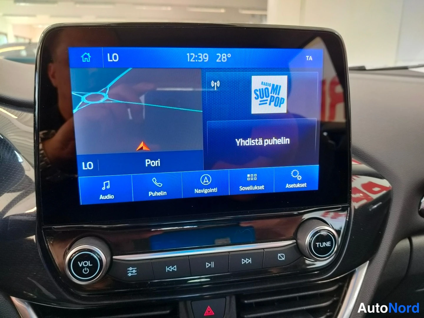 Yksityiskohta: Infotainment-näytön lähikuva: navigaatio kartta näkymällä, radio Radio SuomiPOP ja valikot Audio, Puhelin, Navigointi, Sovellukset, Asetukset.