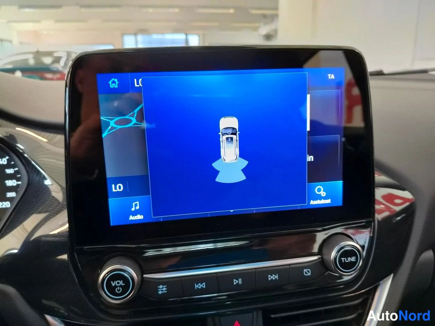 Yksityiskohta: Infotainment-näyttö, jonka päällä avautunut kuvapaneeli autosta ylhäältä katsoen; alareunassa Audio- ja Asetukset-painikkeet.