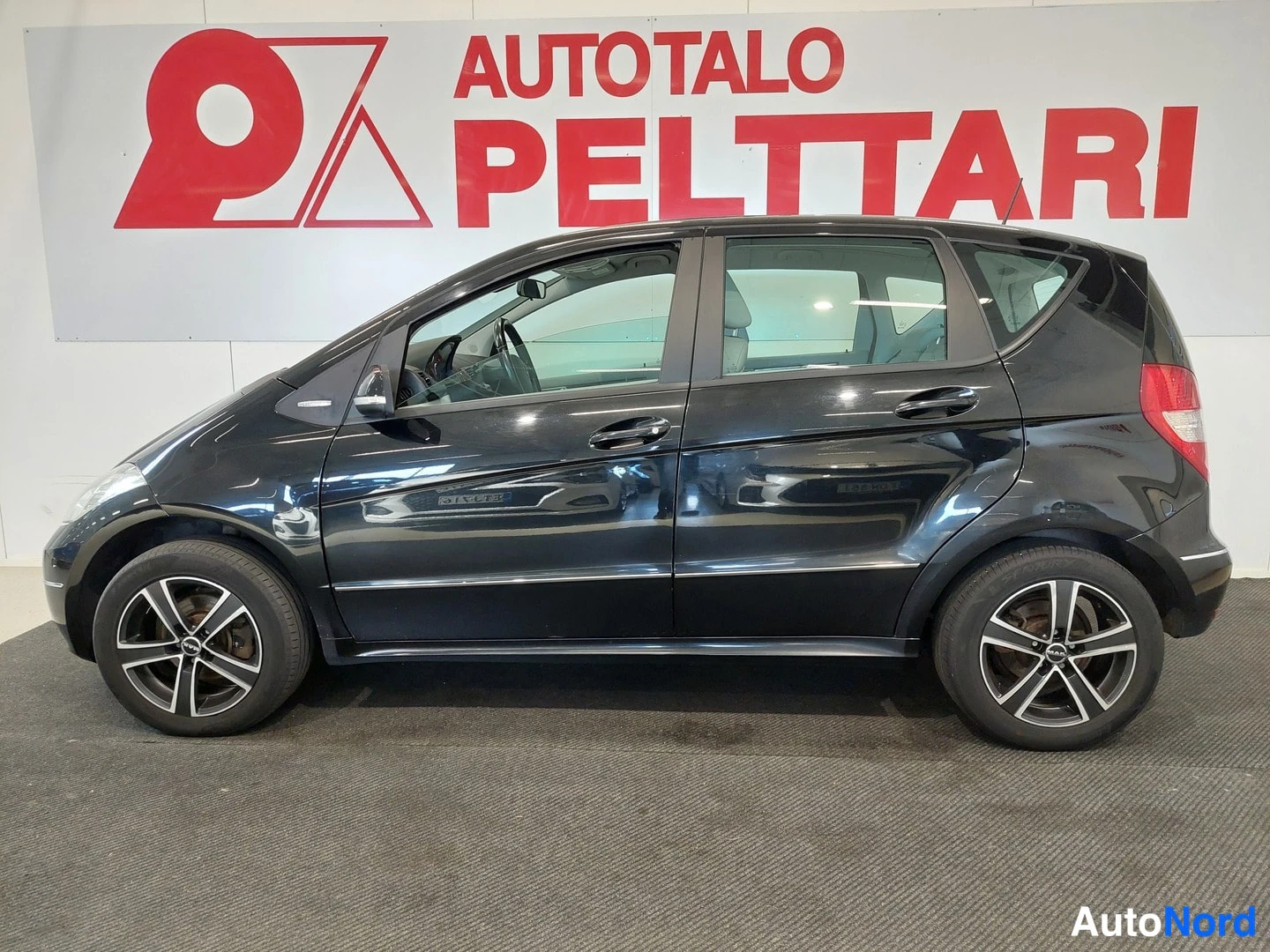 Sivulta: Musta Mercedes-Benz A-sarjan viisiovinen hatchback sivusta oikealta katsottuna, kehysten sijoitus autosalissa.