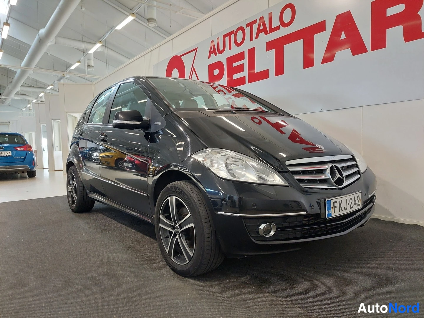 Etuviisto, nokka oikealle: Mustan Mercedes-Benz A-sarjan hatchbackin etukulma vasemmalta, keulakoriste ja rekisteritunnus näkyvissä autosalissa.