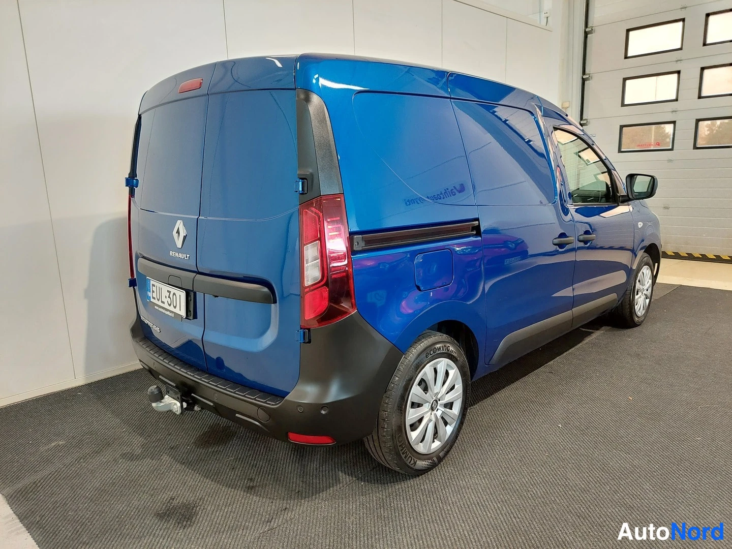 Takaviisto: Sinisen Renault-pakettiauton takaviisto vasemmalta, Renault-merkki, takavalot, rekisterikilpi ja vetokoukku näkyvissä.