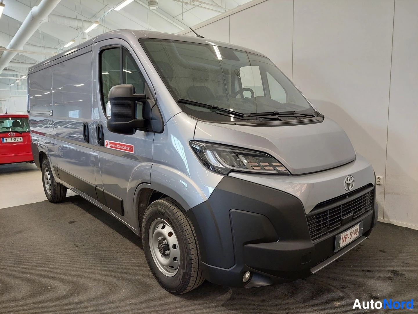 Etuviisto, nokka oikealle: Hopeaharmaa Toyota-pakettiauto kulmasta etupää vasemmalle kuvattuna autosalissa; rekisterikilpi NP-5440, etulokasuojat mustat.