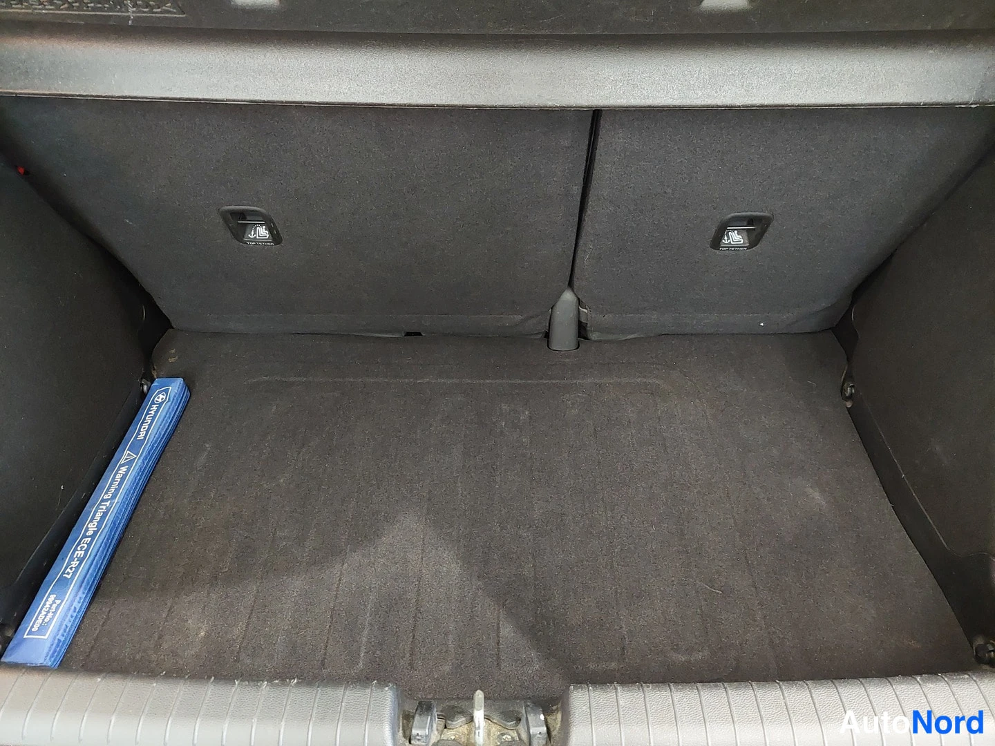 Muu: Tavaratilan sisäkuva: tummat takaselkänojat ja musta matto lattiassa, vasemmalla sininen HYUNDAI varoituskolmio.