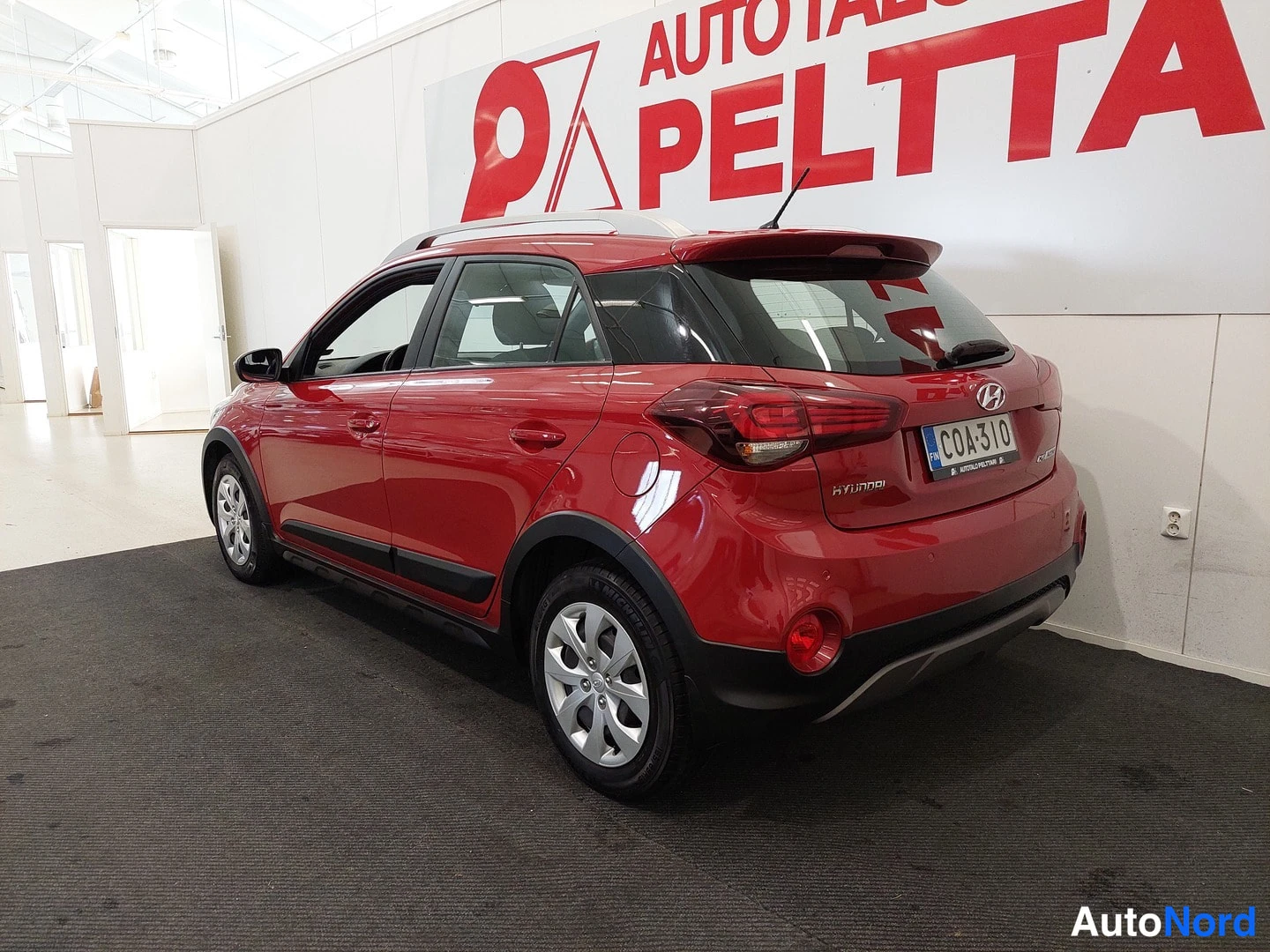 Takaviisto: Punaisen Hyundai-hatchbackin takakulmakuva sisätilassa; takaosat, takaovet ja katetut takavalot näkyvissä, rekisteritunnus.