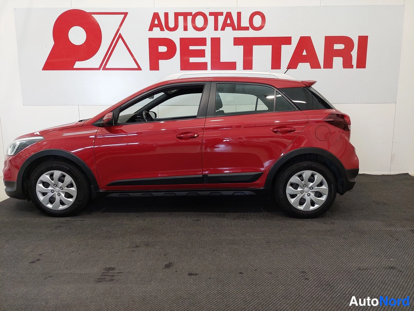 Takaviisto: Punainen Hyundai i20 Active Cross sivusta sisätiloissa, valkoinen katto, tummat muovilistat ja hopeavanteet.