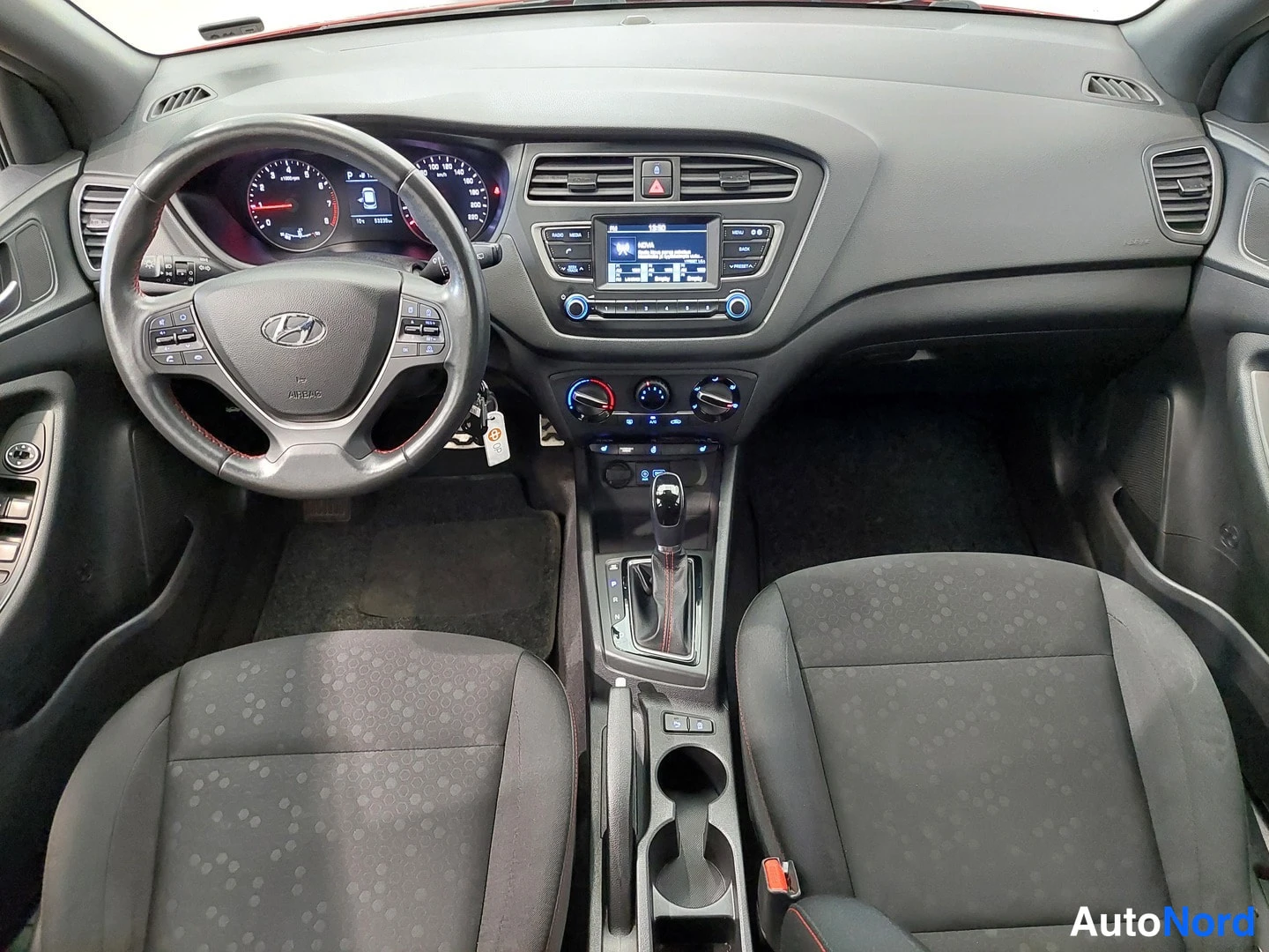 Muu: Hyundai i20:n tumma ohjaamo ylhäältä: ohjauspyörä, mittaristo, radiopaneeli näytöllä ja automaattivaihteen keila.