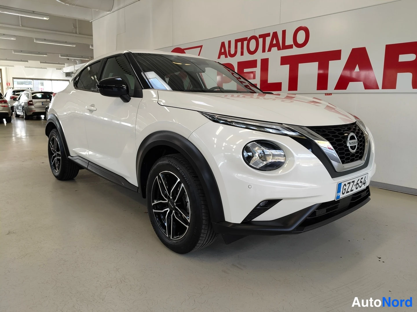 Etuviisto, nokka oikealle: Nissan Juke etuviistosta sisätiloissa; valkoinen kori, etusäleikkö ja rekisteritunnus näkyvissä.