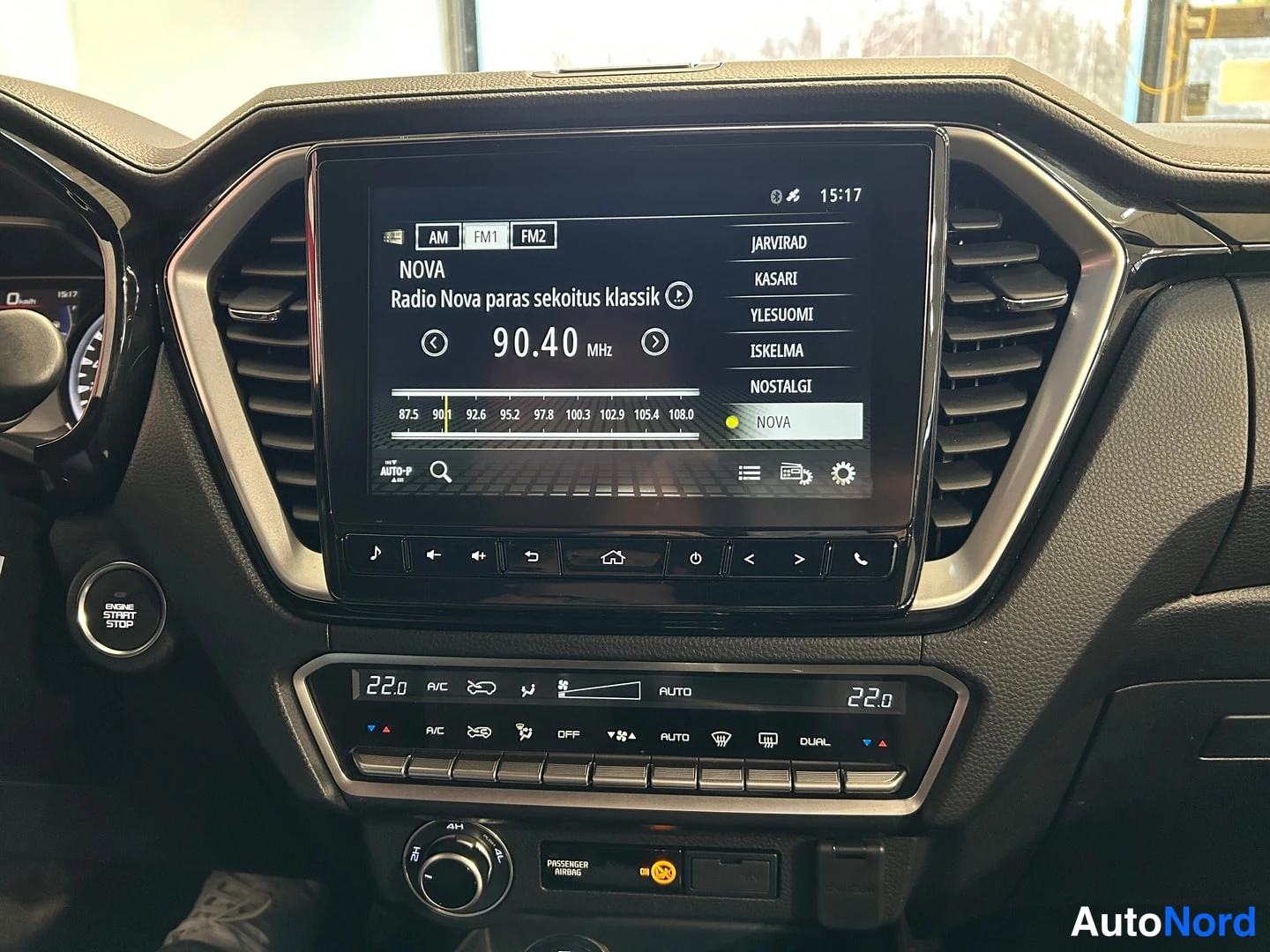 Muu: Kojelaudan infotainment-näyttö näyttää FM-radiota kanavalla Radio Nova 90,40 MHz, alla ilmastointipaneeli ja moottorin käynnistyspainike.