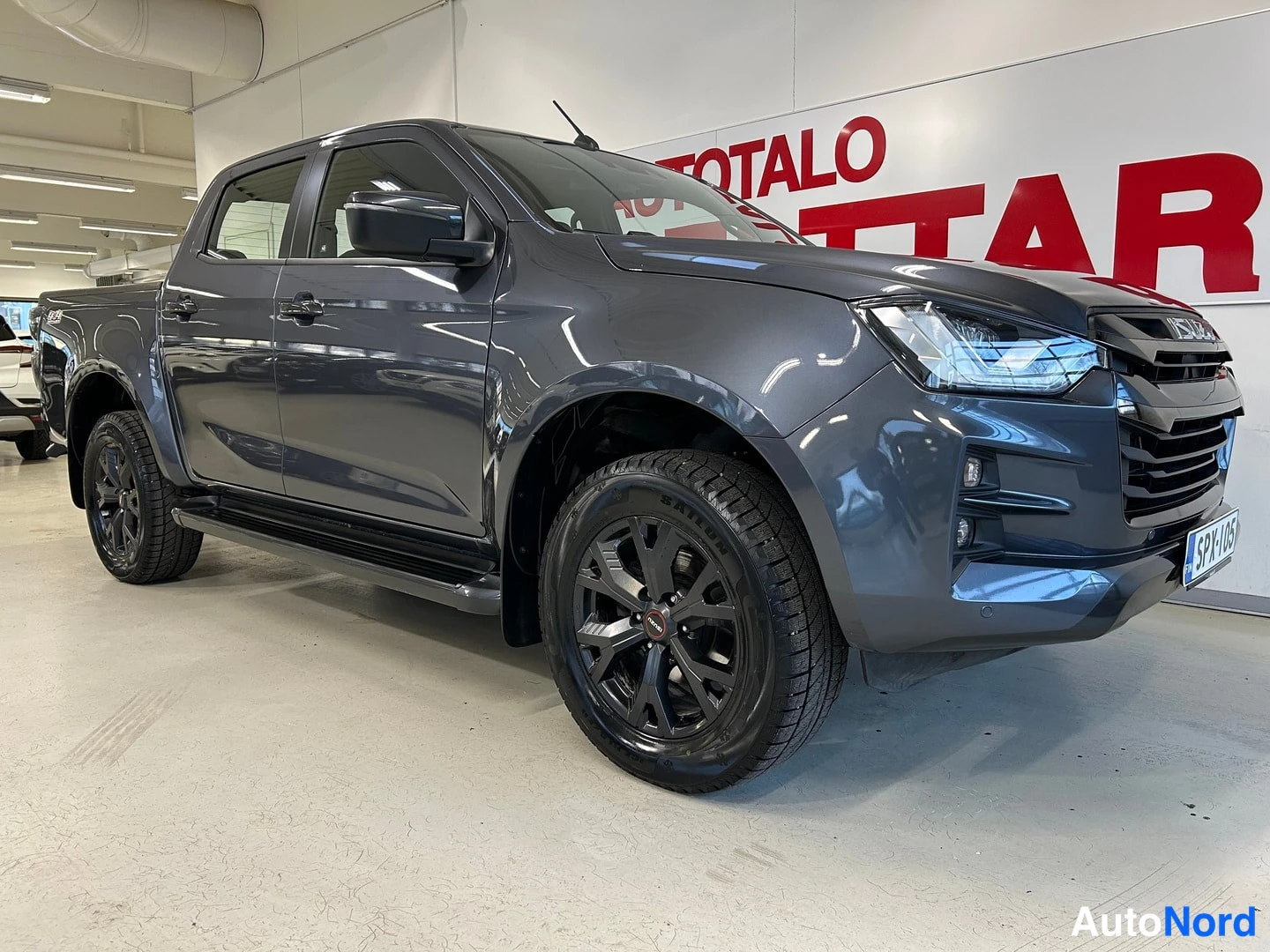 Etuviisto, nokka oikealle: Tummanharmaa avolava-auto keula oikealla viistosti kuvattuna sisätiloissa.