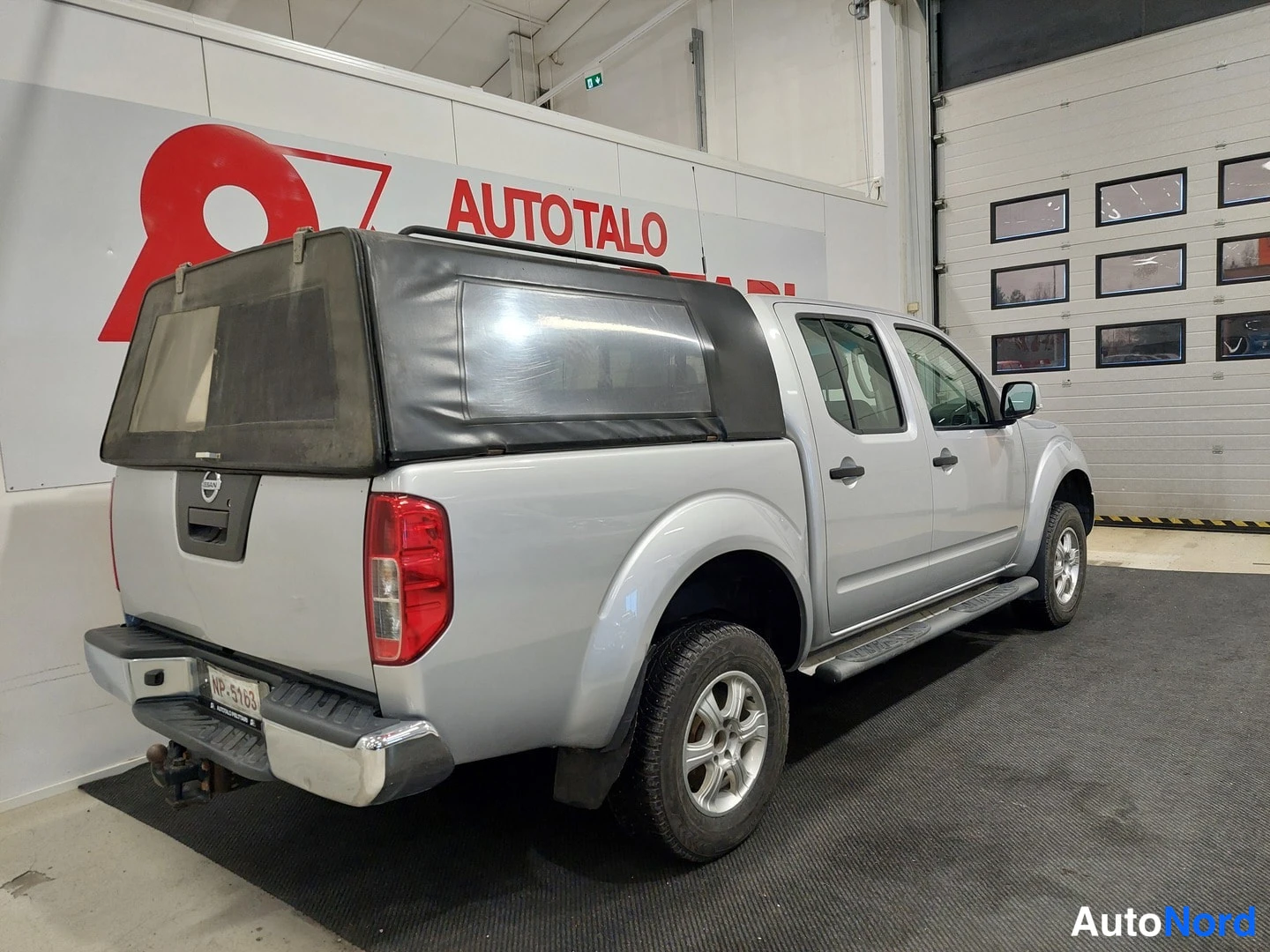 Takaviisto: Nissan Navara Double Cab takaviistosta kuvattuna autotallissa: hopeinen koriväri, tumma kuormasuoja, sivulaitahalkaisu ja viisiovinen ohjaamo näkyvät.