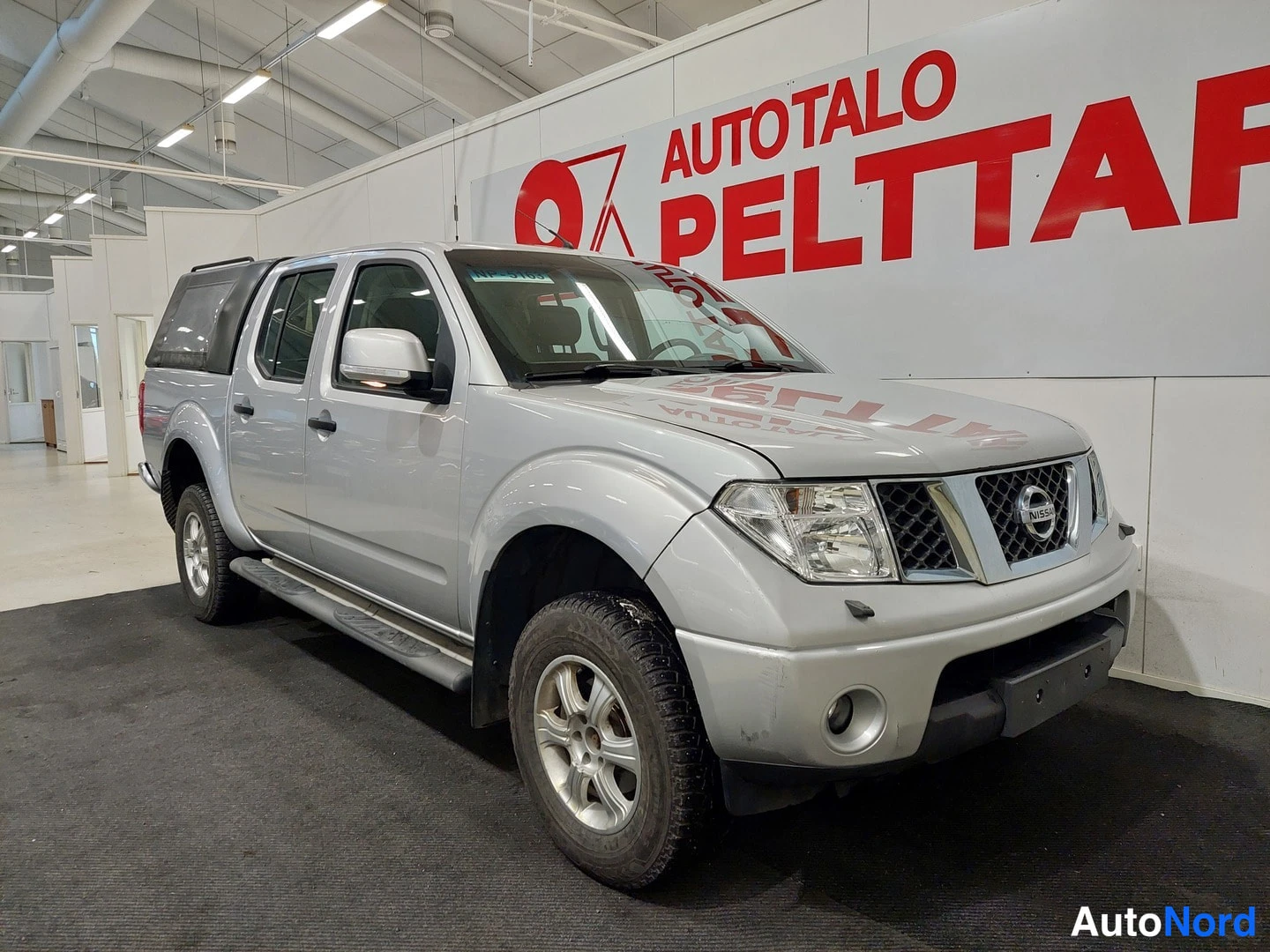 Etuviisto, nokka oikealle: Hopeanvärinen Nissan Navara -avolava-auto etuviistöstä kuvattuna; keulagrilli ja kylki näkyvät samassa kuvassa sisätiloissa.