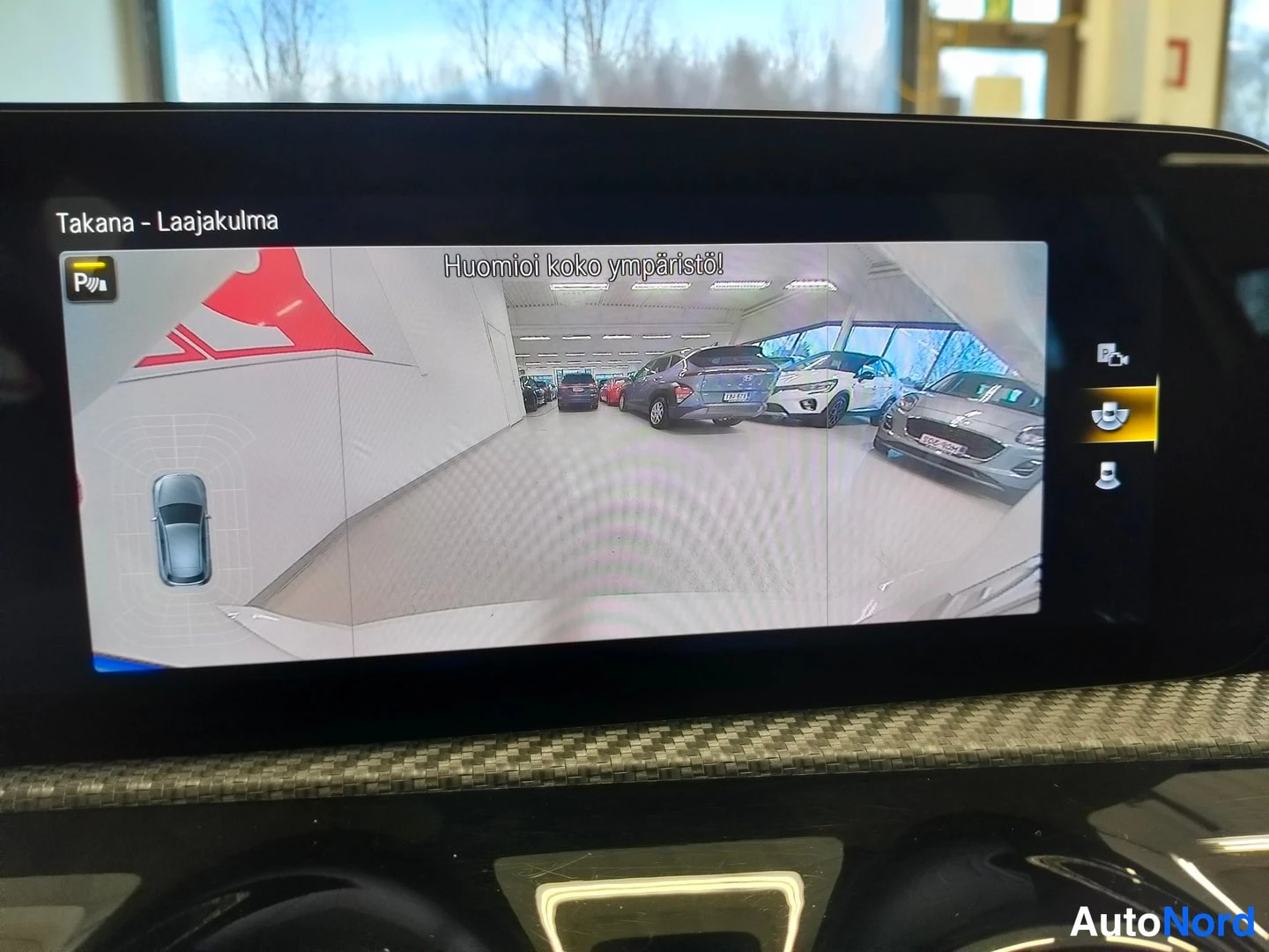Muu: Kojelaudan näytöllä näkyy laajakulmainen peruutuskameran kuva showroom-tilasta, jossa useita autoja, ja otsikkoteksti 'Takana - Laajakulma'.