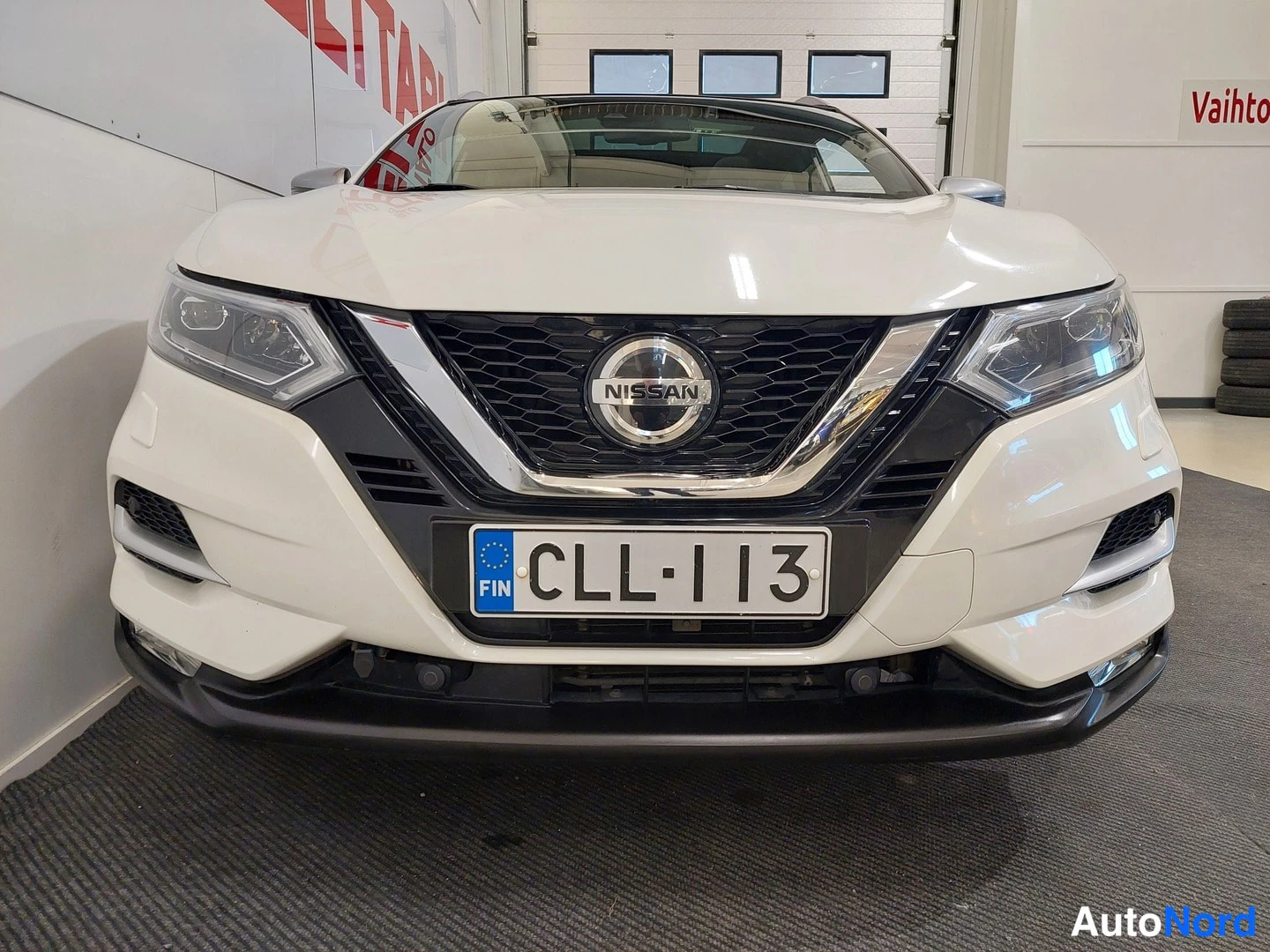 Edestä: Auton etupään tarkennus: Nissan-merkki sälekuidissa, -rekisterikilpi ja sumuvalot.