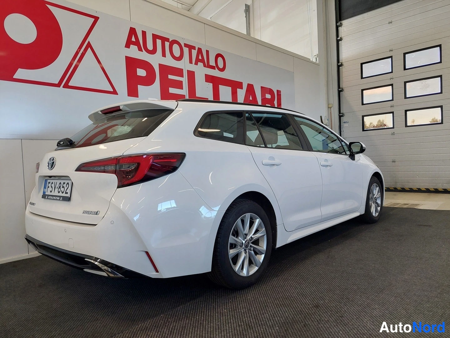 Takaviisto: Valkoinen Toyota Corolla -farmari takaa ja oikealta sivusta nähtynä sisätiloissa, takavalot ja rekisterikilpi näkyvät.