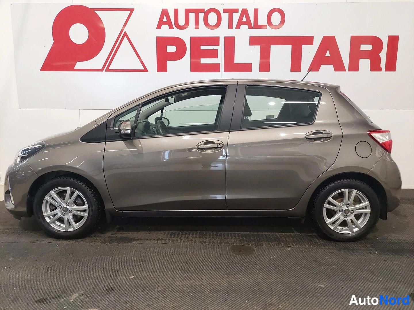 Sivulta: Auton vasen kylki sivulta kuvattuna; ruskeaharmaa viisiovinen hatchback, hopeavanteet, myyntihallin seinä taustalla.