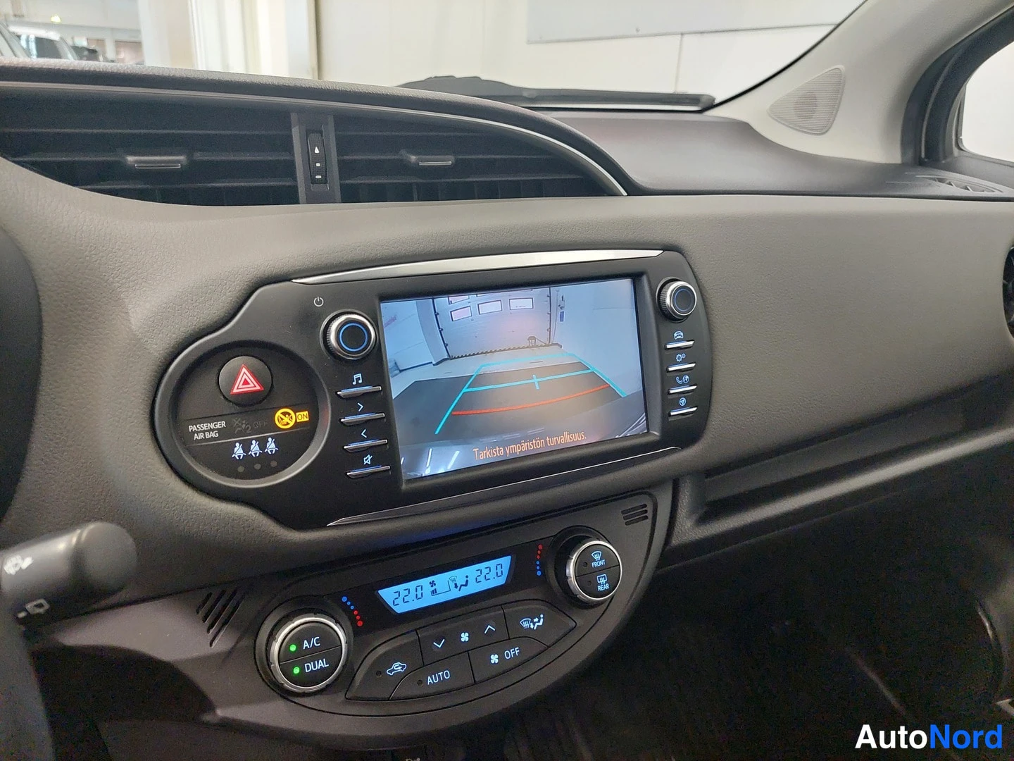 Sivulta: Toyota Yarisin kojelauta: infotainment-näyttö näyttää peruutuskameran kuvan tekstillä 'Tarkista ympäristön turvallisuus', alla kaksipaikkainen ilmastointi A/C D