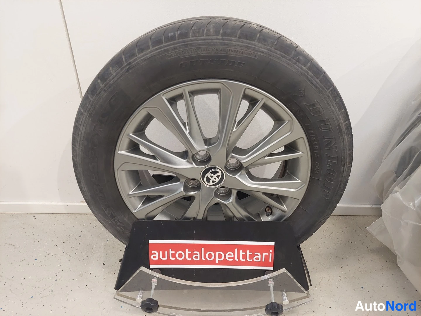 Yksityiskohta: Dunlop-kesärengas Toyotan hopeisella alumiinvanteella esitteishyllyssä, taustalla valkoinen seinä ja 'autotalopelttari'-kyltti.