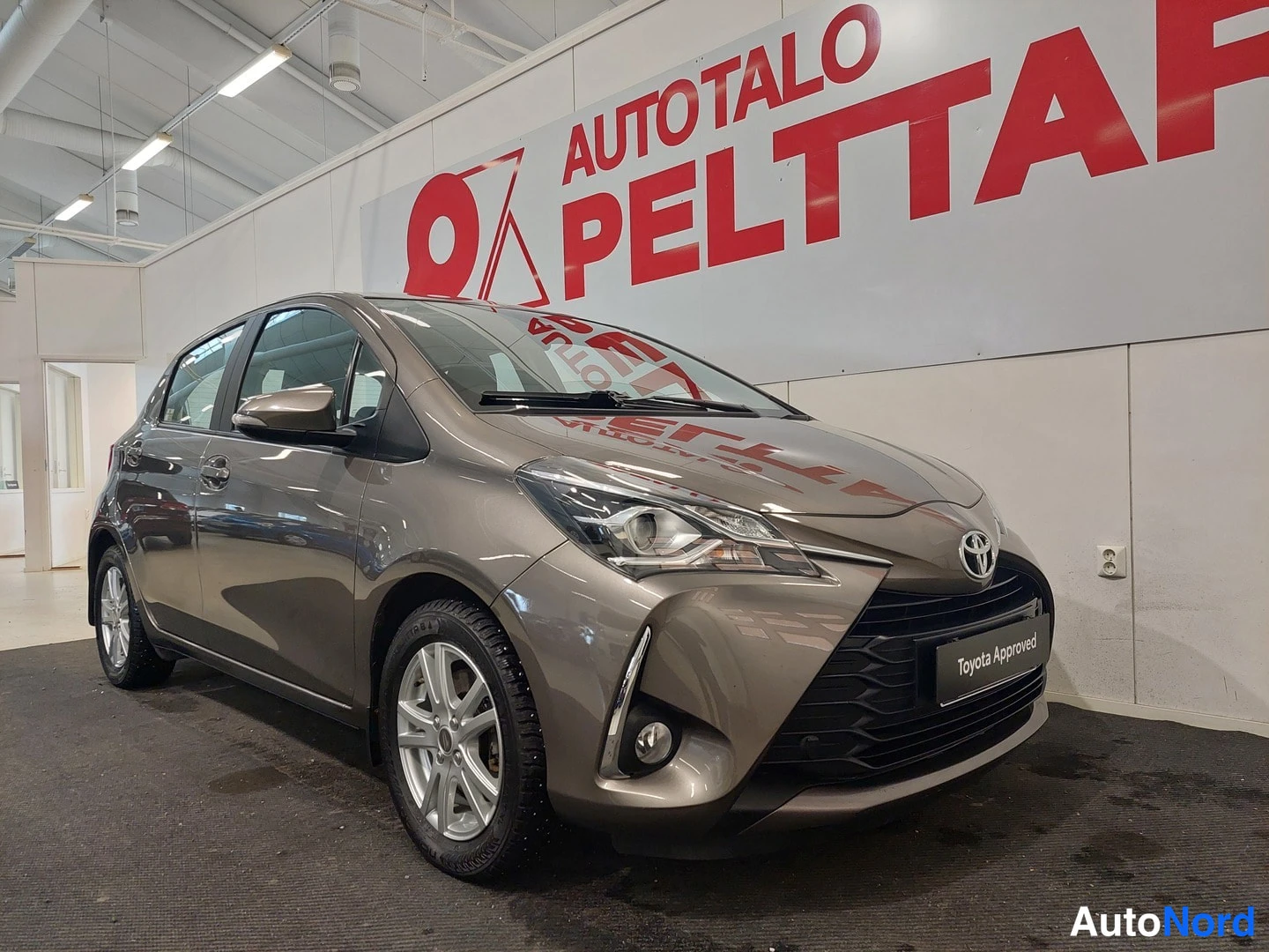 Etuviisto, nokka oikealle: Ruskeanharmaan Toyota Yaris -henkilöauton etukulma näkyy sisätiloissa, konepellin kirkkaus heijastaa mainoslogon.