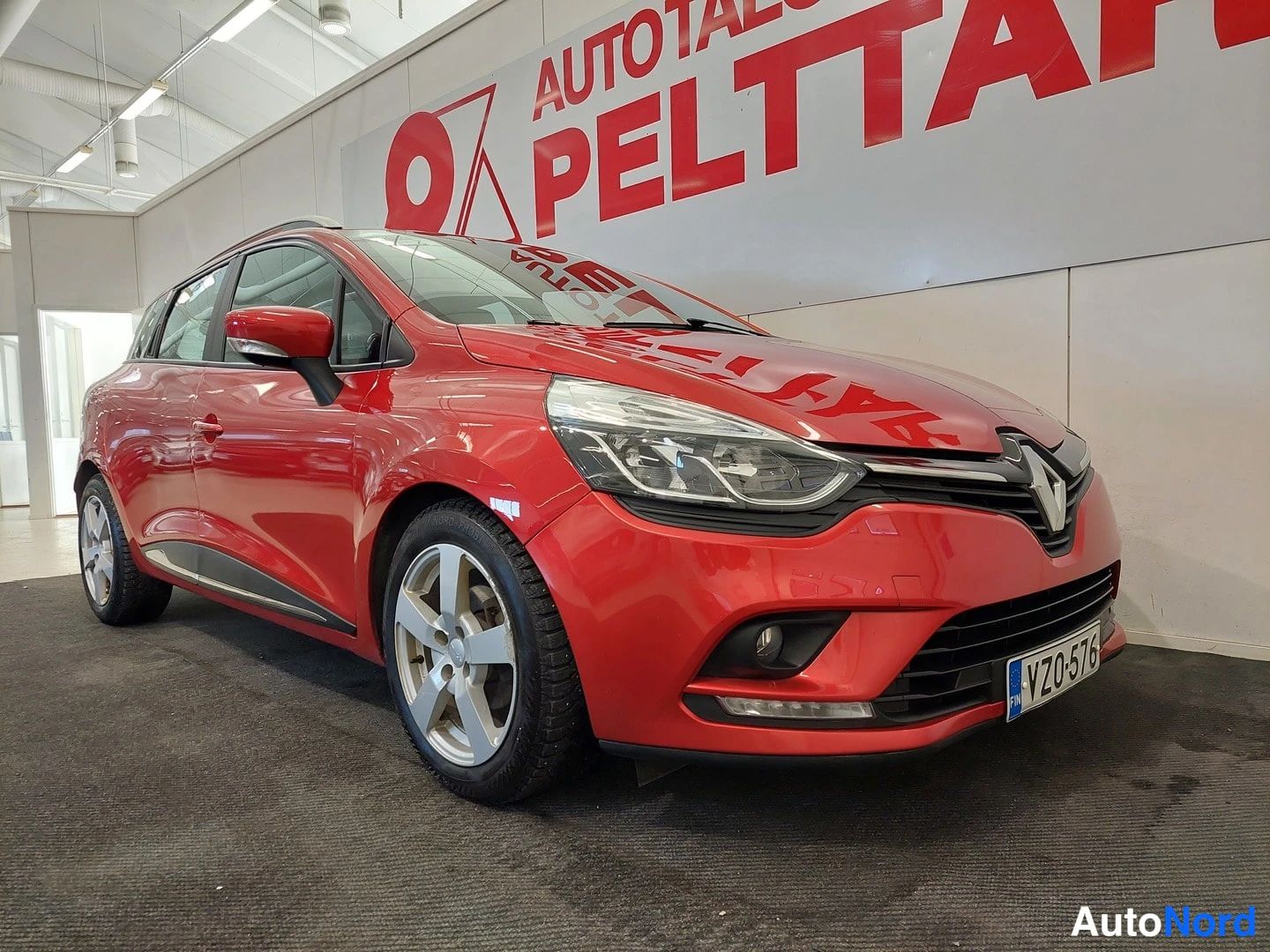 Etuviisto, nokka oikealle: Punainen Renault Clio Sport Tourer etuosaltaan kolmannesvasemmasta kuvattuna sisätiloissa, etupuskuri ja rekisterikilpi näkyvissä.