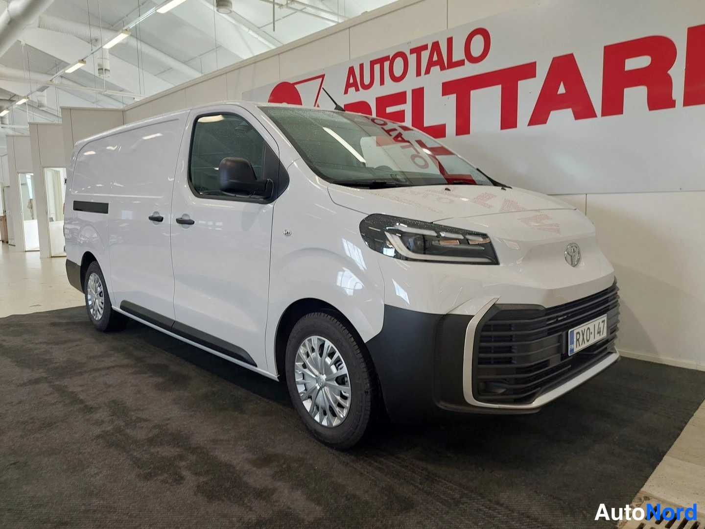 Etuviisto, nokka oikealle: Valkoinen Toyota Proace -pakettiauto nähtynä etuviistöstä autosalin lattialla, rekisterikilpi.