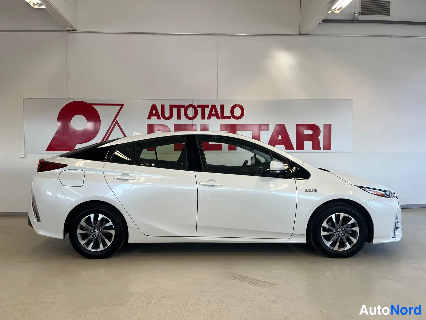 Takaviisto: Valkoinen Toyota Prius Plug-In -henkilöauto sivulta takaviistoon kuvattuna sisätiloissa, perän ja oikean sivun ollessa näkyvissä.