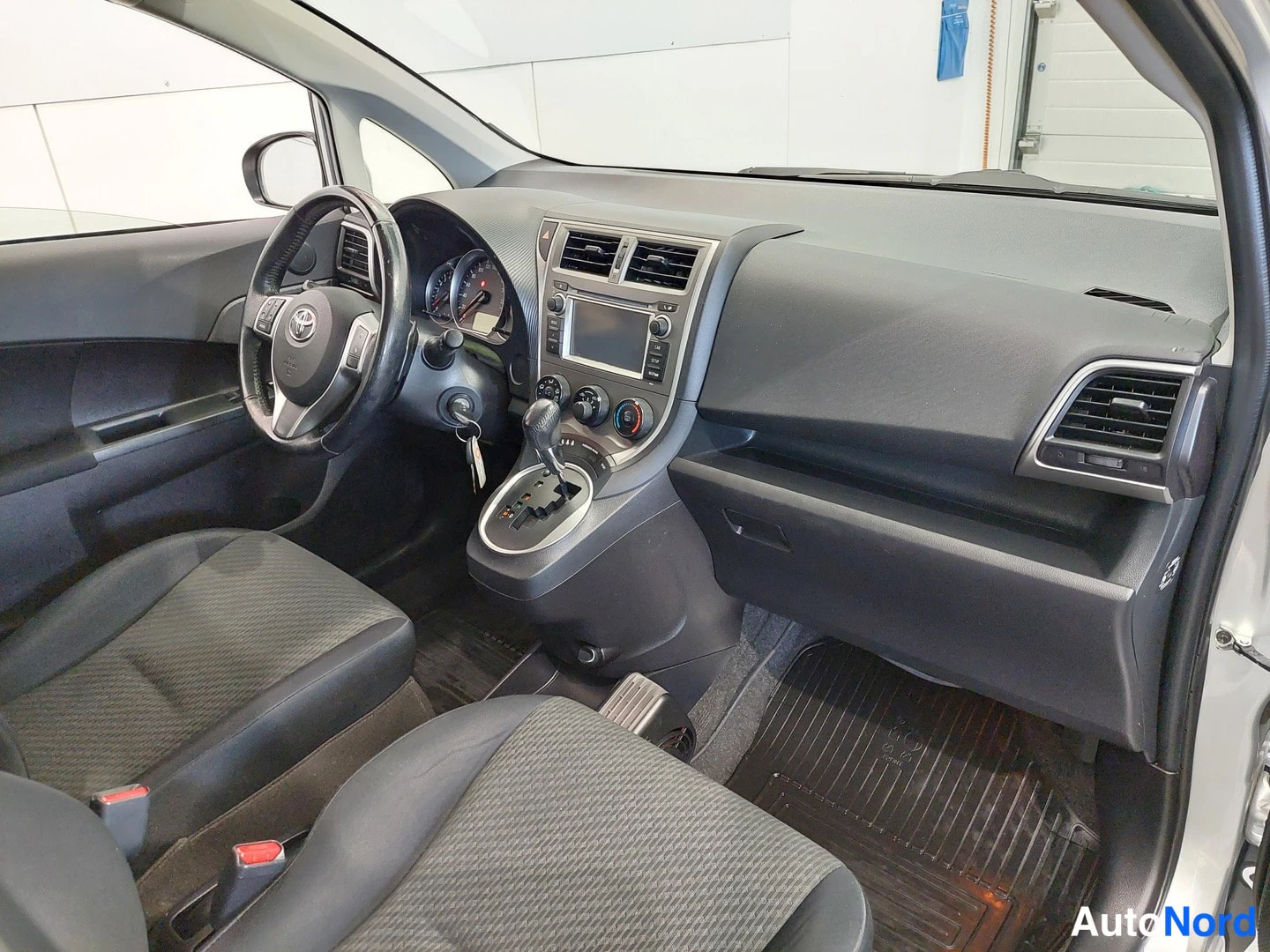 Edestä: Auton sisätila: tumma kojelauta, Toyota-ohjauspyörä, automaattivaihteen valitsin, keskikonsolin näyttö ja kaksi etuistuinta.