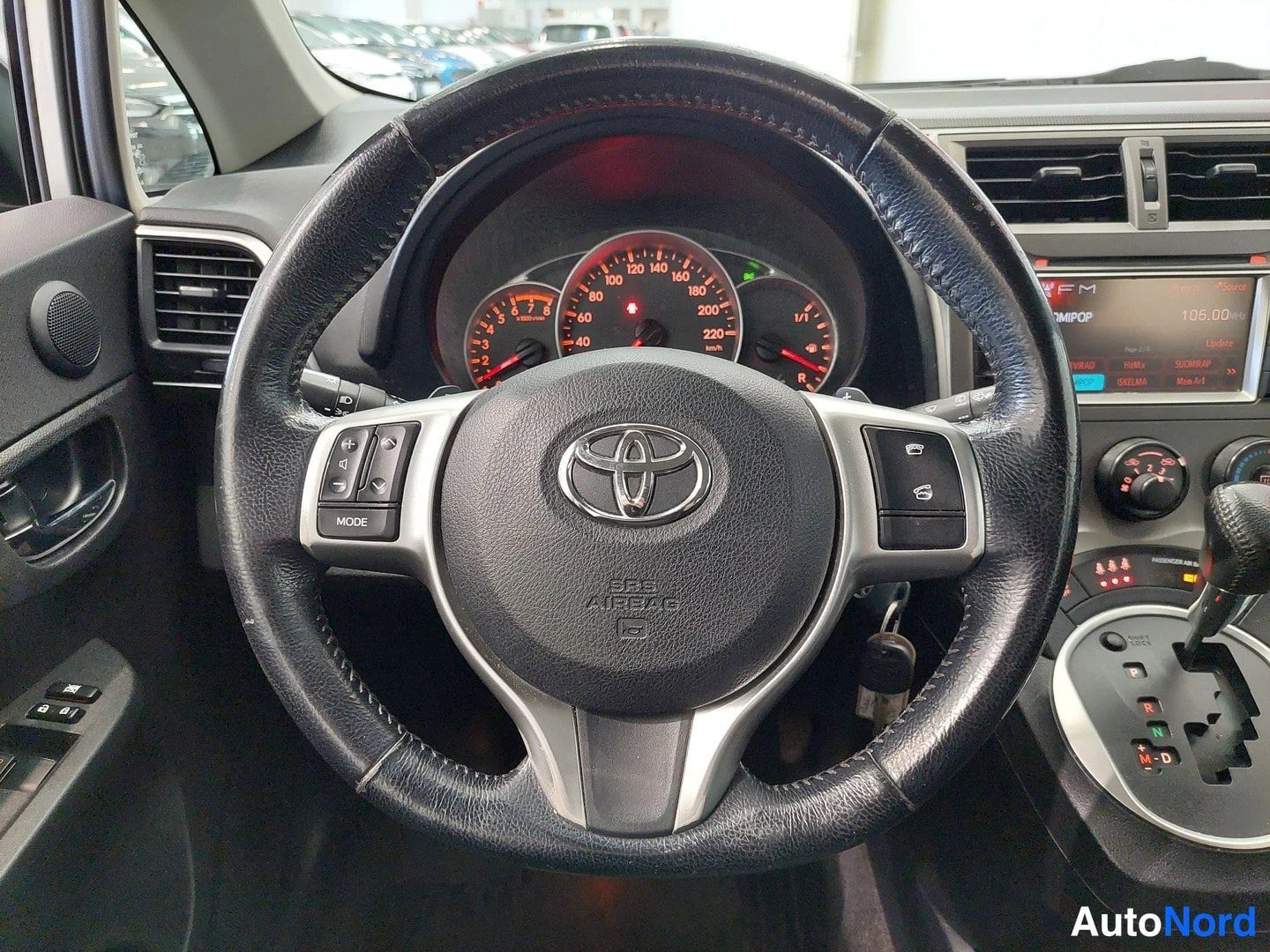 Muu: Ohjauspyörä Toyota-logolla, SRS AIRBAG -teksti ja monitoiminapit vasemmalla ja oikealla; taustalla mittaristo, radiopaneeli (FM 106.00) ja automaattivaihteiston
