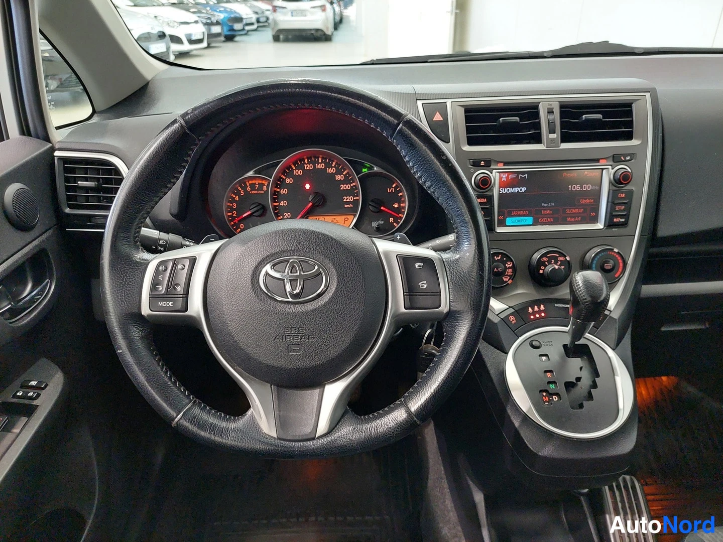 Muu: Ohjauspyörä ja kojelauta lähikuvassa: Toyota-merkki, mittaristo, radio ja automaattivaihteiston valitsin näkyvissä.