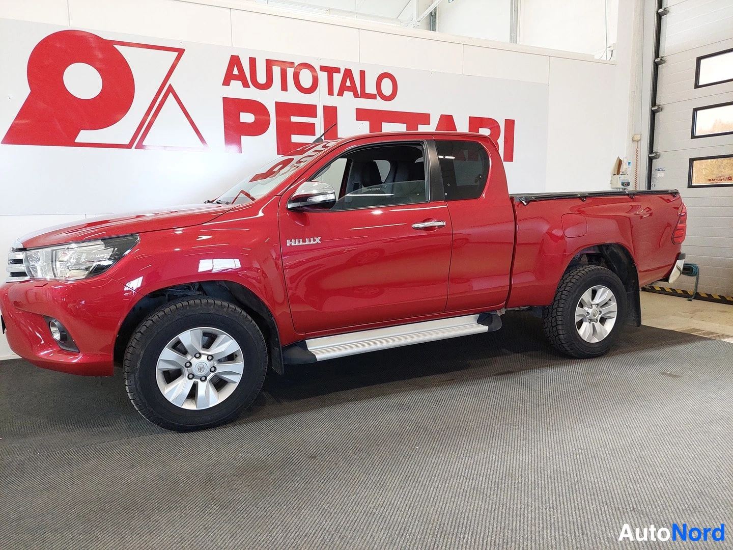 Etuviisto, nokka vasemmalle: Punainen kaksiovinen Toyota Hilux -avolava sivulta ja etukulmasta; KULUX-tarra ovesta luettavissa, mukana sivukorokkeet ja takaseinän suojaverho.