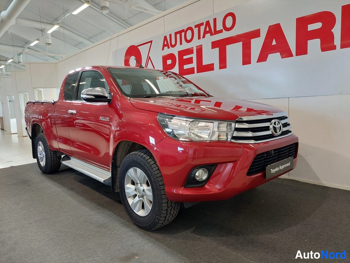 Etuviisto, nokka oikealle: Punainen avolava-auto etuviistosta kuvattuna sisätiloissa; keulagridia, Toyota-merkki ja 'Toyota Approved' -kyltti näkyvissä.