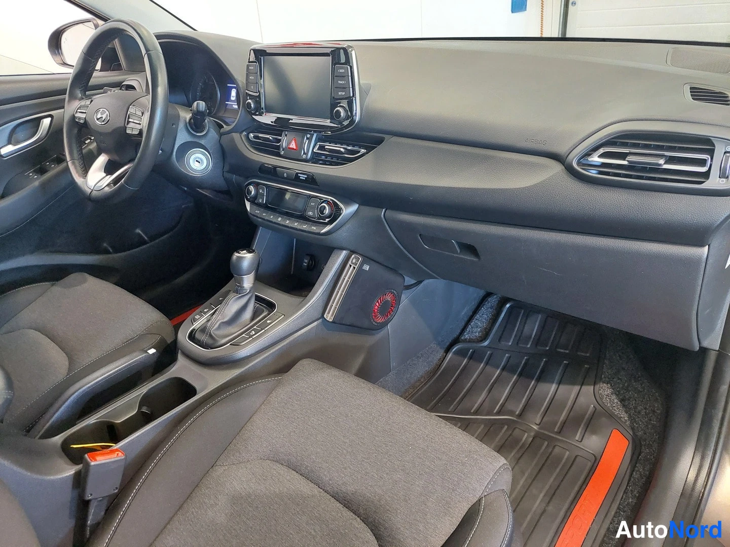 Muu: Etumatkustustila ja kojelauta Hyundai i30:ssä, manuaali- vaihtautomaatti ja mattonaat näkyvissä.