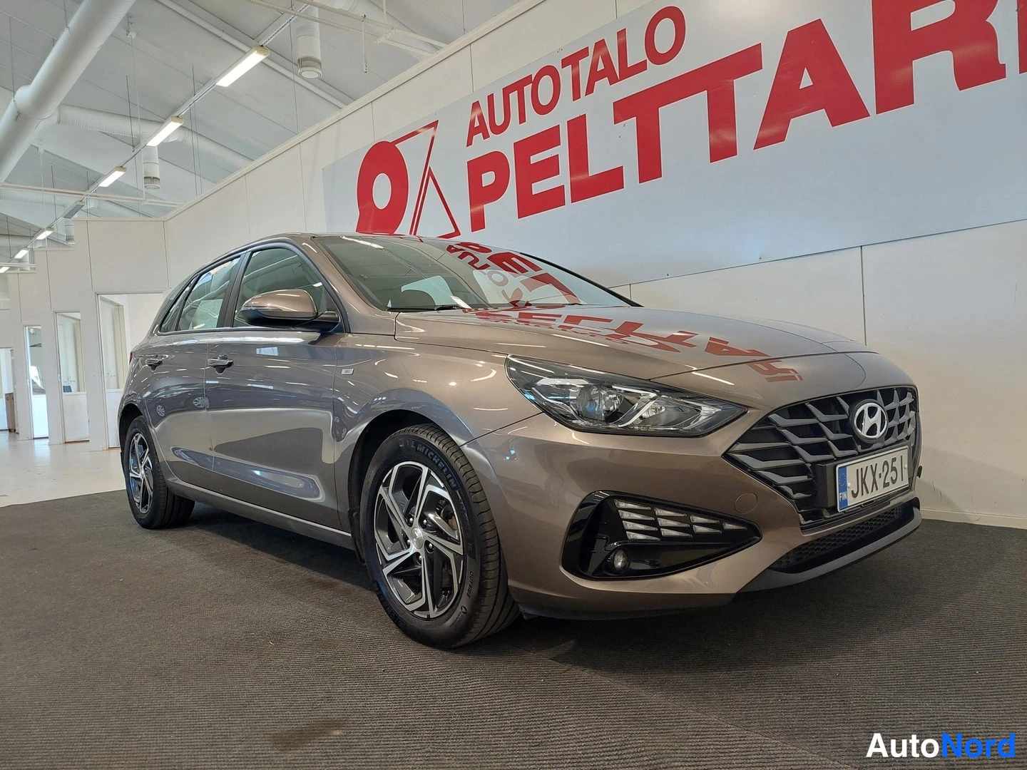 Etuviisto, nokka oikealle: Hyundai i30 -viistoperä etuviistolta oikealta, hopeanruskea, Autovalo Pelttari -mainostaulun edessä.
