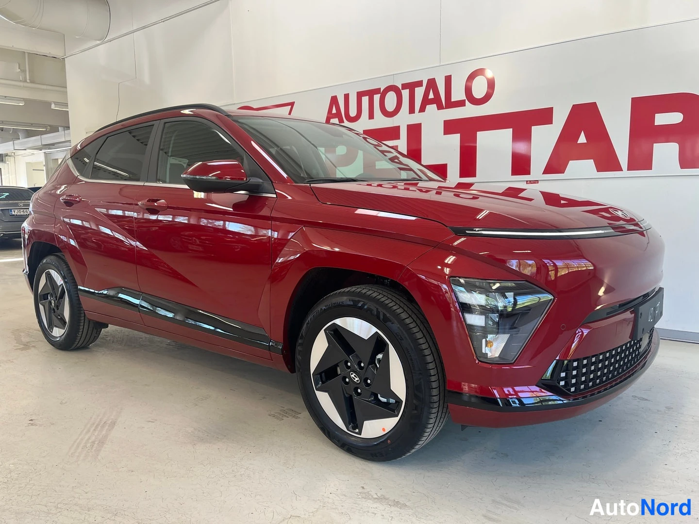 Etuviisto, nokka oikealle: Punainen Hyundai Kona Electric -maasturin etukulmanäkymä sisätiloissa, keulakerta ja vasen sivu näkyvissä.