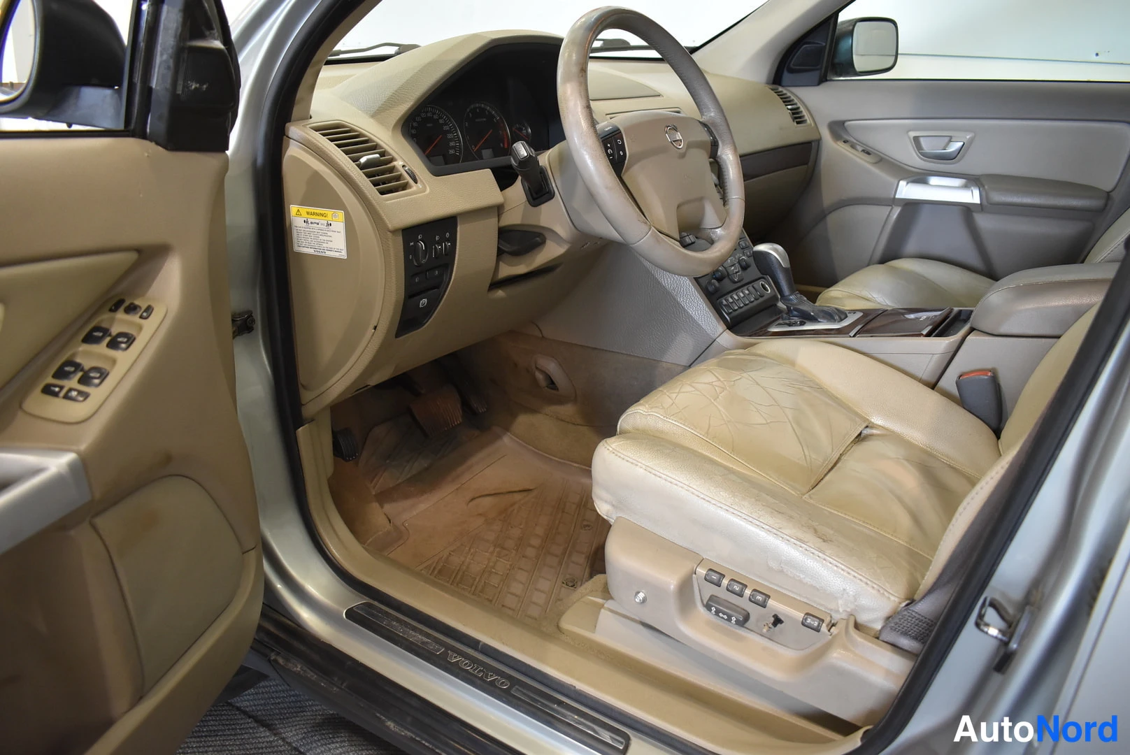 Muu: Volvo XC90:n sisätilat kuljettajan oven kautta: beige/punertava nahkasisusta, ohjauspyörä, automaattivalitsin ja penkkien säätönapit.