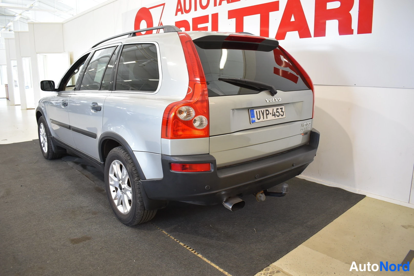 Takaviisto: Hopeanharmaa Volvo XC90 -maasturin takanurkkakuva sisätilassa; rekisterikilpi, kattokaiteet ja takatuli- ja peruutusvalot näkyvissä.