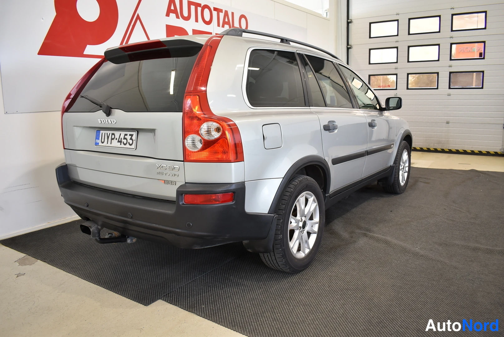 Takaviisto: Hopeinen Volvo XC90 perästä ja oikeasta sivusta sisätiloissa, takamoottori- ja rekkyteline näkyvät.