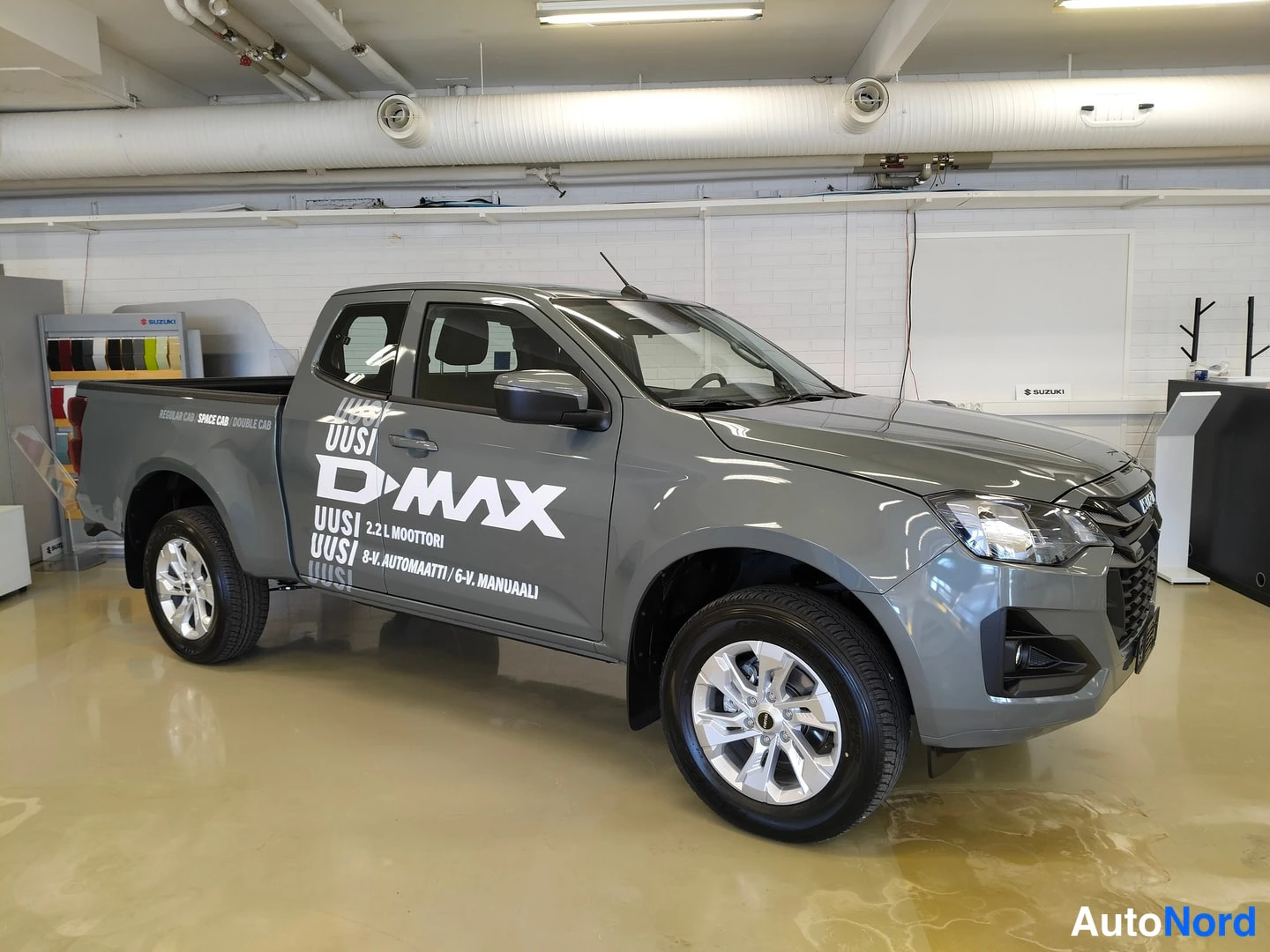 Etuviisto, nokka oikealle: Harmaa Isuzu D-Max avolava-auto sisätiloissa, etukulma oikealta, kyljessä D-MAX-kuvat
