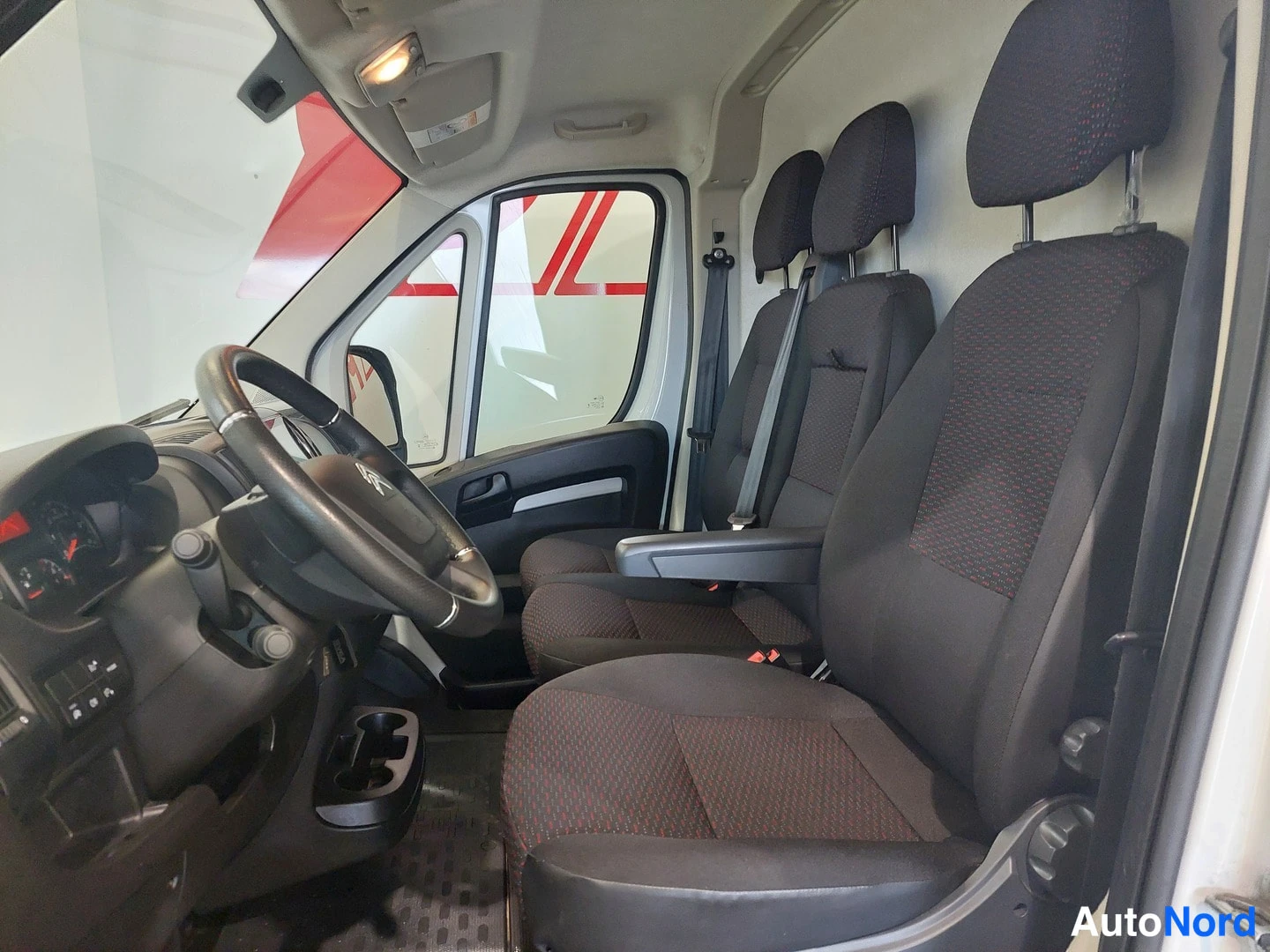 Muu: Pakettiauton ohjaamo: kuljettajan istuin, kaksi matkustajainstuinta ja Citroën-ohjauspyörä tummassa kankaassa.