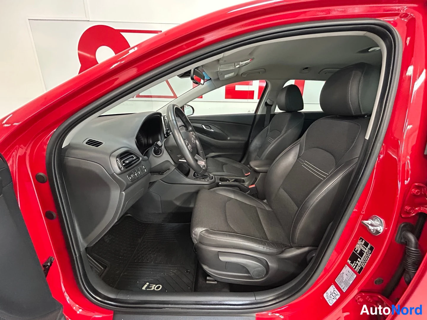 Muu: Avoimen etuoven näkymä Hyundai i30:n sisätilaan: tummat etuistuimet, ohjauspyörä, keskikonsoli ja tekstiilityynystetty lattiamatisto, jossa lukee i30.