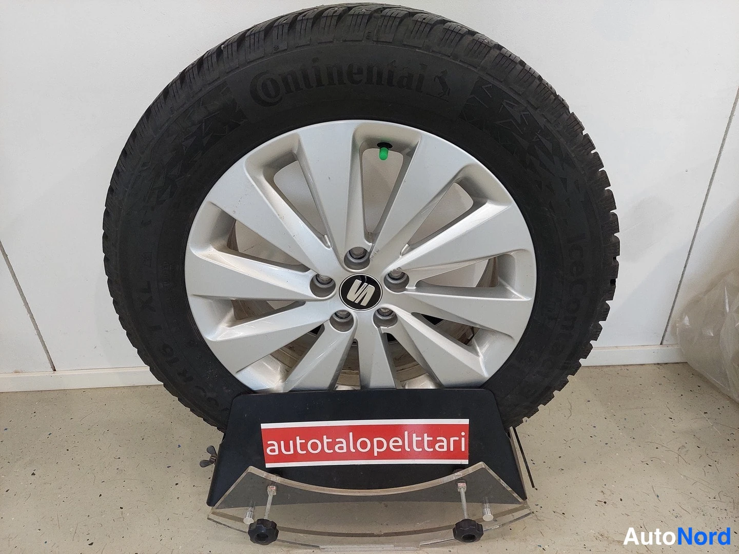 Yksityiskohta: Kokonainen talvipyörä esillä telineessä, jossa on punainen teksti autotalopelttari; hopeinen Seat-vanne ja musta Continental-rengas.
