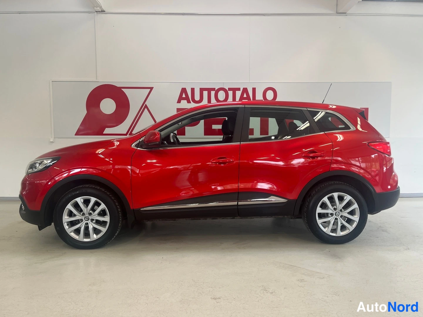 Sivulta: Punaisen tila-auton sivuprofiili vasemmasta etukulmasta, hopeiset vanteet ja kromikoriste ovien alla valkoisella seinällä myyntitaulu.