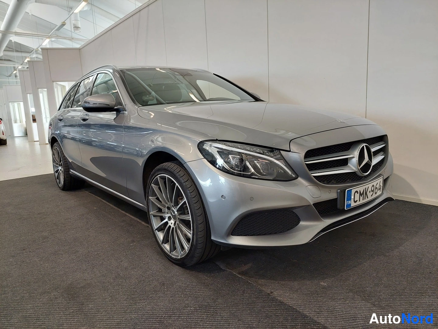 Etuviisto, nokka oikealle: Hopeanharmaa Mercedes-Benz farmari etuviistosta oikealta kuvattuna sisätiloissa, rekisterikilpi.